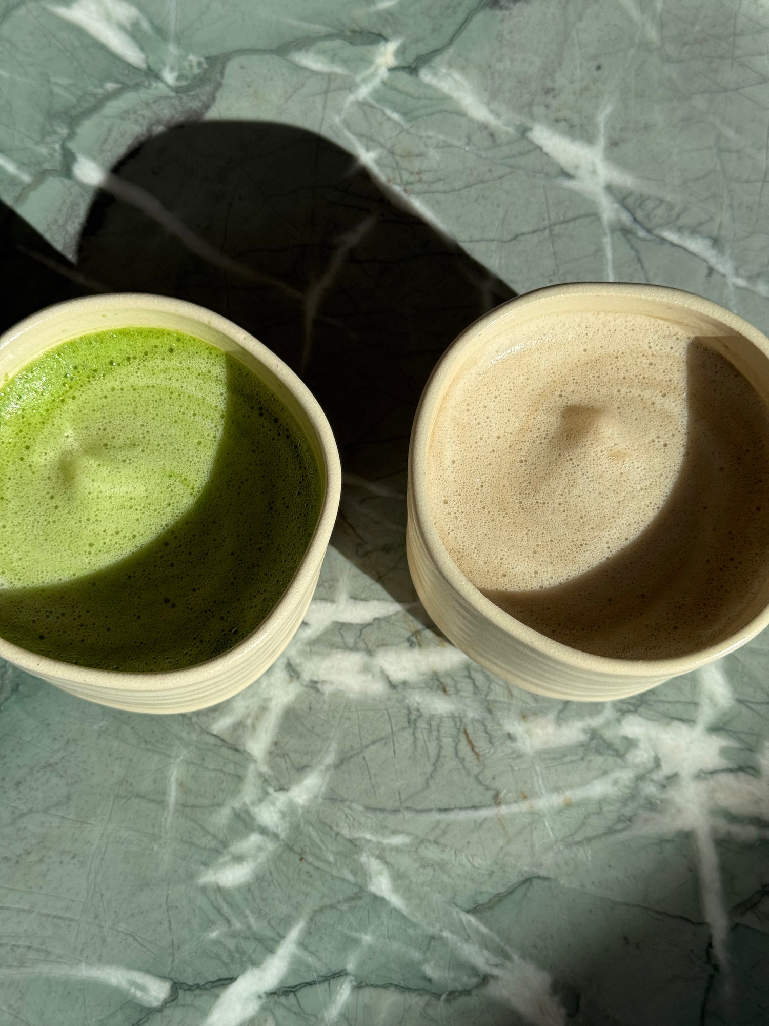 Matcha und Hojicha Latte in zwei Keramikschalen auf Marmortisch im Sonnenlicht