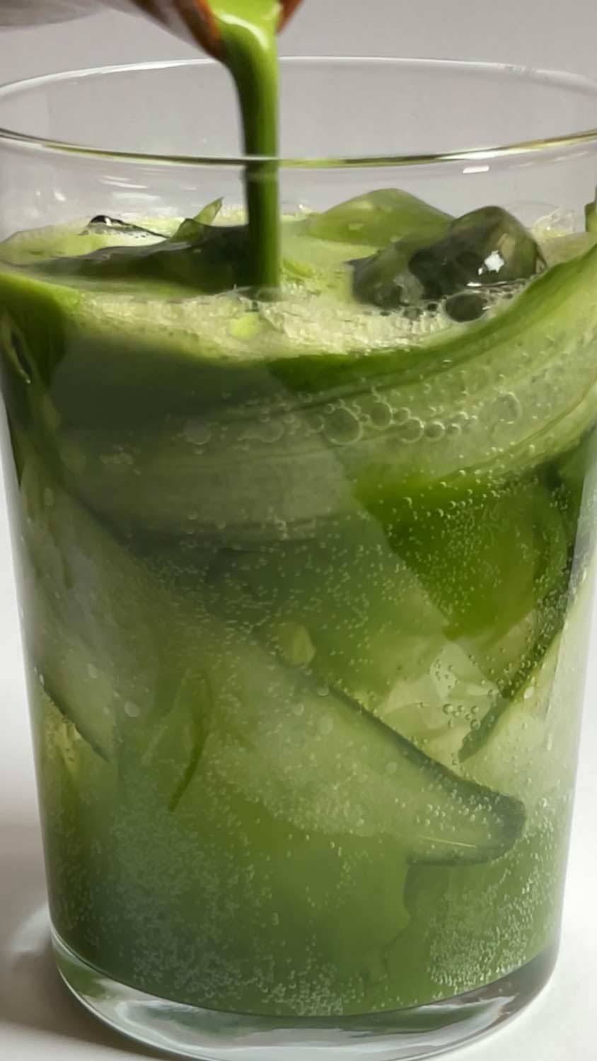 Cucumber Kombucha Matcha im Glas mit Eiswürfeln und Gurkenscheiben während grüner Matcha hineingegossen wird