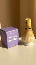 Matcha Besen aus hellem Bambus auf weißem Whisk Holder neben lila Verpackung mit Aufschrift Whisk Holder Pearl White von Health Bar, warmes Sonnenlicht und minimalistischer Hintergrund für stilvolles Matcha Zubehör