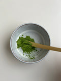 Feines Bio Matcha Pulver in hellgrauer Keramikschale mit Bambuslöffel, Draufsicht auf leuchtend grünes Matcha Pulver vor weißem Hintergrund für die traditionelle Zubereitung von Matcha Tee