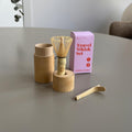 Travel Whisk Set von Health Bar mit Bambus Chasen, passender Aufbewahrungsdose aus Bambus, Chasen Halter und Bambuslöffel vor rosa Verpackung auf Tisch in natürlichem Licht