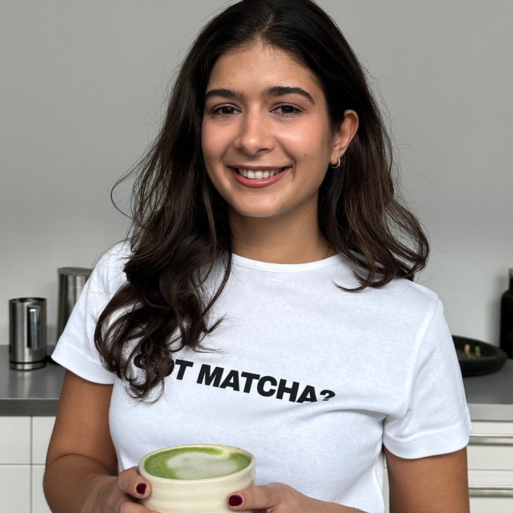 Frau mit weißem Got Matcha? Shirt hält eine Tasse Matcha Latte und lächelt in die Kamera