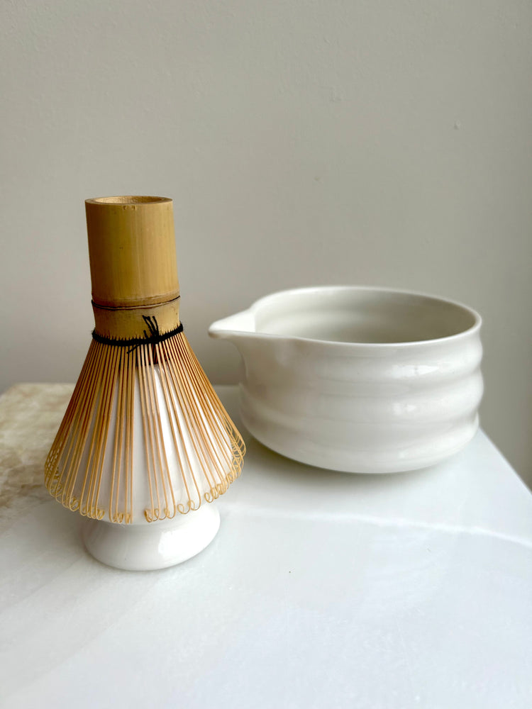 Matcha Zubehör Set mit Bambus Chasen Besen und weißer Keramik Mixing Bowl mit Ausguss, minimalistisch fotografiert auf hellem Untergrund vor neutraler Wand, natürliche Lichtstimmung für traditionelle Matcha Zubereitung